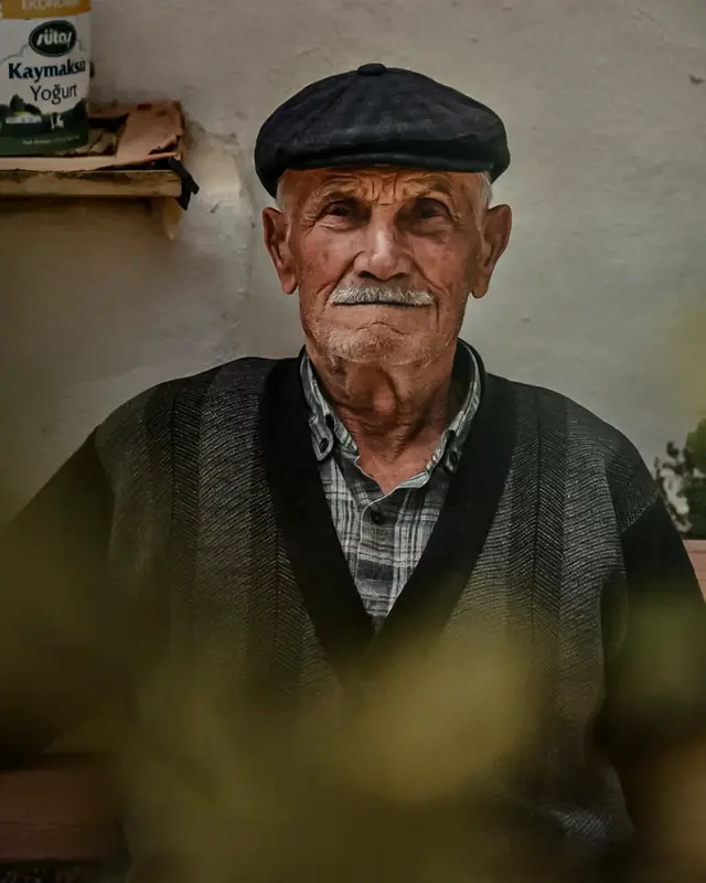 An older man in a flat cap sitting calmly outdoors behind soft greenery